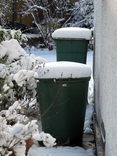 Twee groene regentonnen met een laag sneeuw tegen de huismuur in de winter
