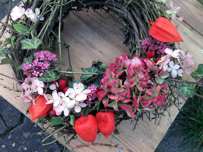 Zelfgebonden herfstkrans met roze bloemen, oranjerode lampionnen en groene bladeren op een plank